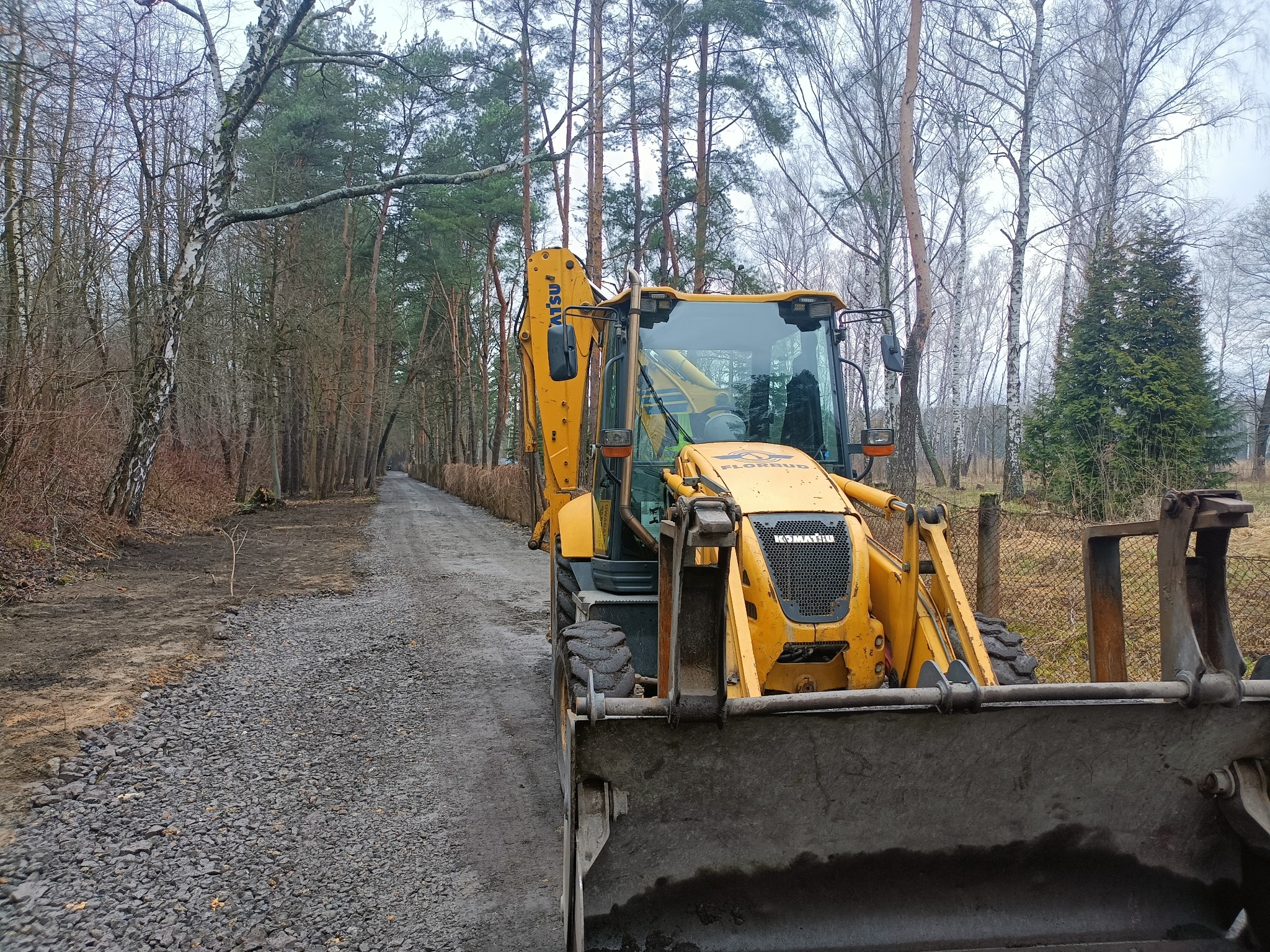 Żółta koparko-ładowarka Komatsu na żwirowej drodze w lesie, widoczne świeżo wysypane kamienie.