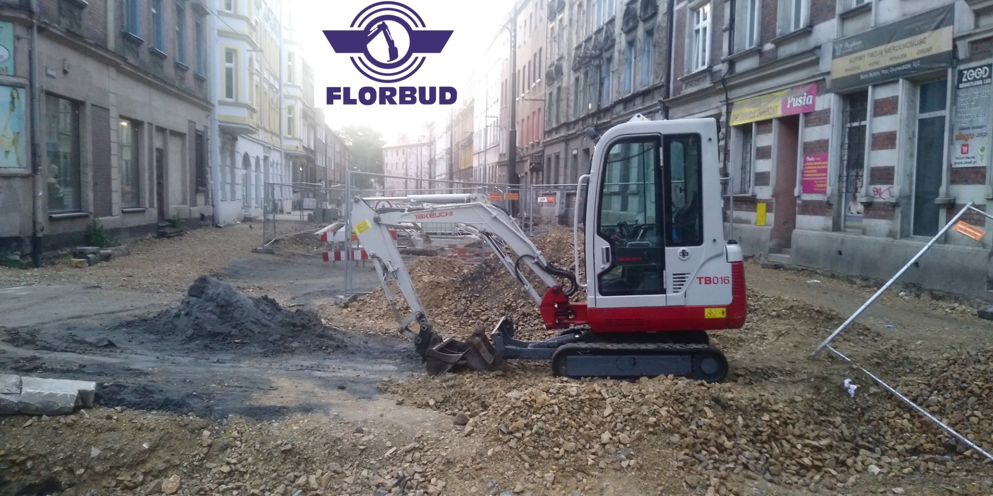 Mini koparka Takeuchi TB016 na tle miejskiej ulicy w trakcie prac ziemnych, z widocznym stosem ziemi i zabezpieczeniami, logo firmy Florbud w górnej części kadru.