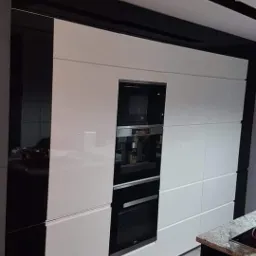 Biała zabudowa kuchenna z wbudowanym piekarnikiem, ekspresem do kawy i mikrofalówką, czarne elementy dekoracyjne sufitu, widoczny fragment blatu z kamienia.