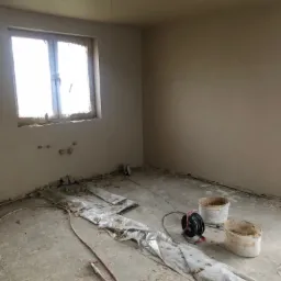 Pokój w trakcie prac wykończeniowych: świeżo położona gładź na ścianach, zabezpieczona folią podłoga, puste wiadra po masie i zwinięty kabel elektryczny.