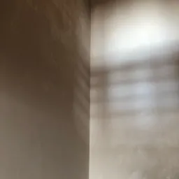 Ściana wewnętrzna w rogu pomieszczenia, częściowo pokryta świeżo nałożoną, nierówną warstwą szarej masy szpachlowej, z widocznymi śladami po narzędziach i kablem wystającym z sufitu.