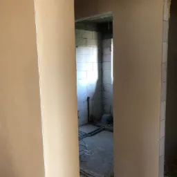 Wnętrze w trakcie prac wykończeniowych: ściany po nałożeniu tynku maszynowego, widoczne instalacje elektryczne i hydrauliczne, ściany z bloczków betonowych w pomieszczeniu obok.