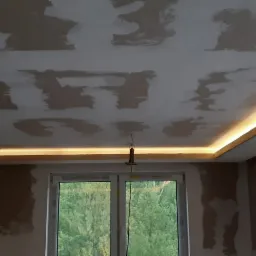 Sufit po szpachlowaniu, z zamontowanym podświetleniem LED wzdłuż krawędzi, widoczne niedoskonałości i nierówności na powierzchni, okno z widokiem na zieleń na zewnątrz.