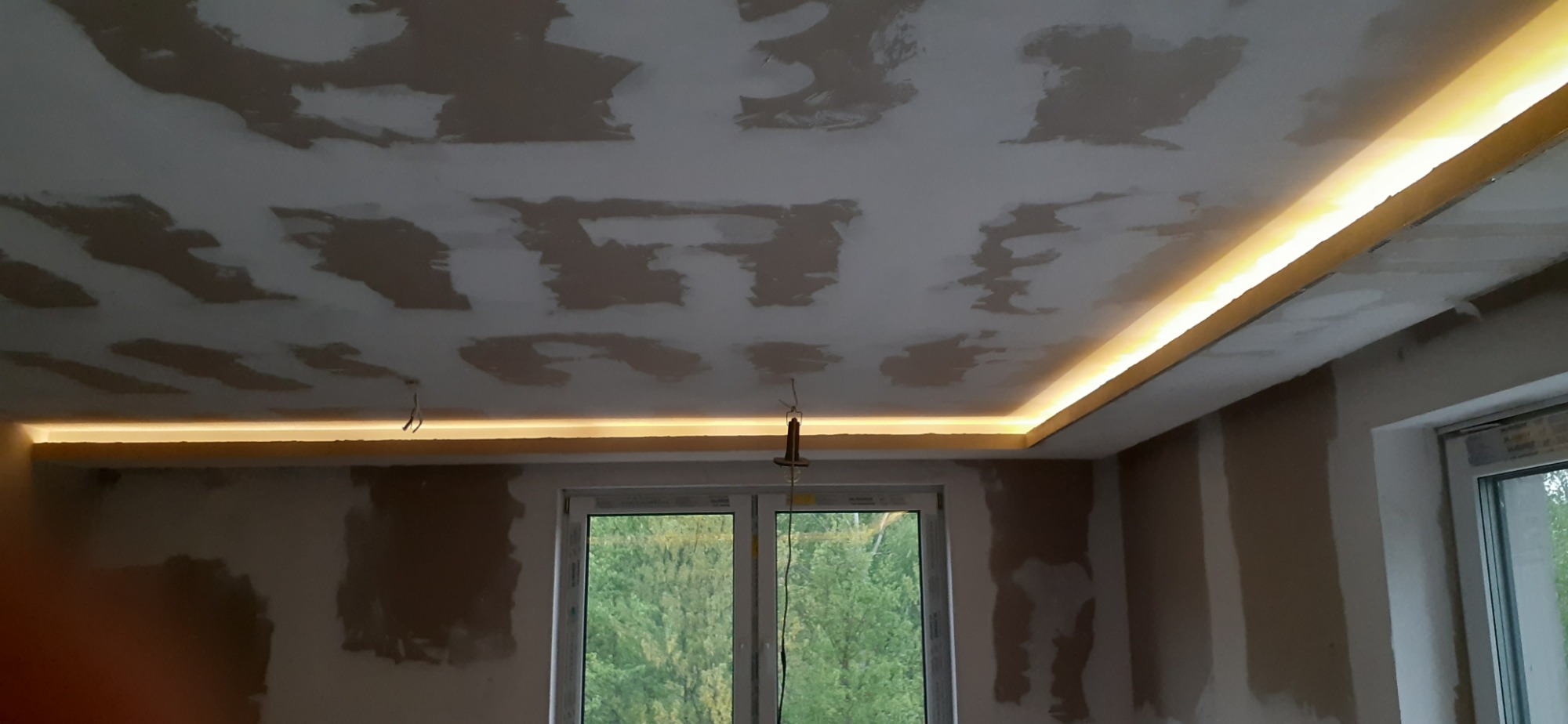 Sufit po szpachlowaniu, z zamontowanym podświetleniem LED wzdłuż krawędzi, widoczne niedoskonałości i nierówności na powierzchni, okno z widokiem na zieleń na zewnątrz.