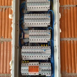 Rozdzielnica elektryczna zamontowana w ścianie z cegły, zabezpieczona pianką montażową, widoczne liczne bezpieczniki i przewody w kolorze niebieskim, żółtym i czarnym.