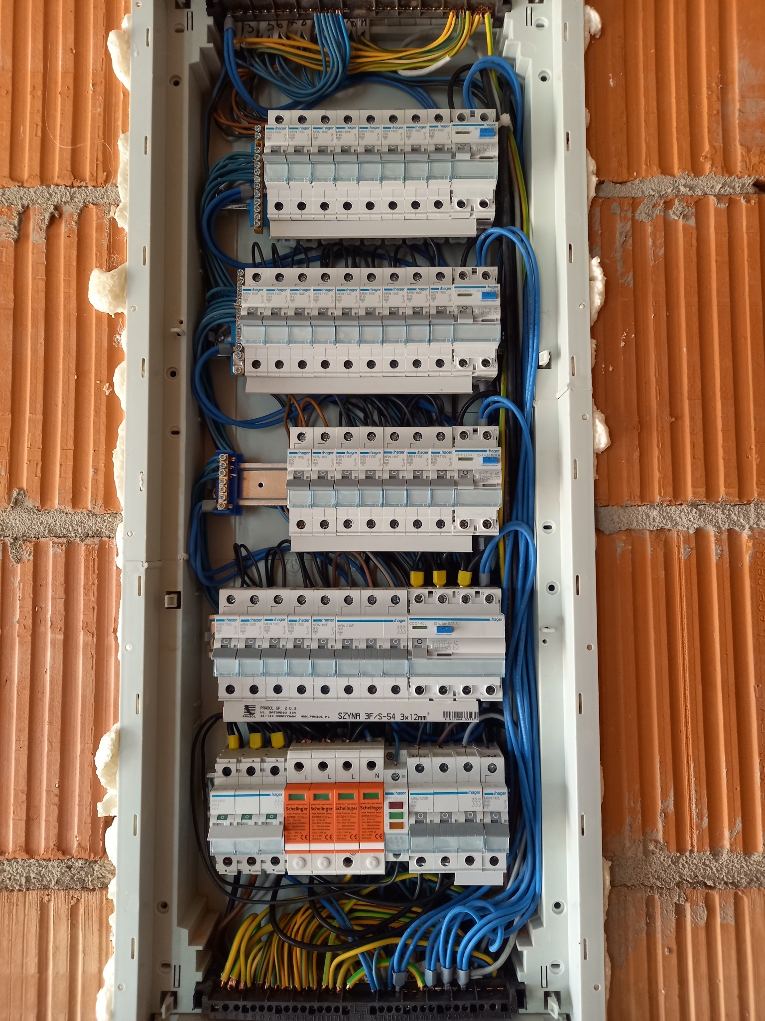 Rozdzielnica elektryczna zamontowana w ścianie z cegły, zabezpieczona pianką montażową, widoczne liczne bezpieczniki i przewody w kolorze niebieskim, żółtym i czarnym.