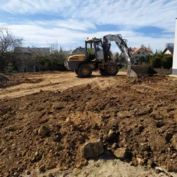 Żółto-szara koparka Mecalac MX100 na placu budowy, widoczny świeżo wyrównany teren z brązową ziemią, w tle dom jednorodzinny i błękitne niebo z chmurami.