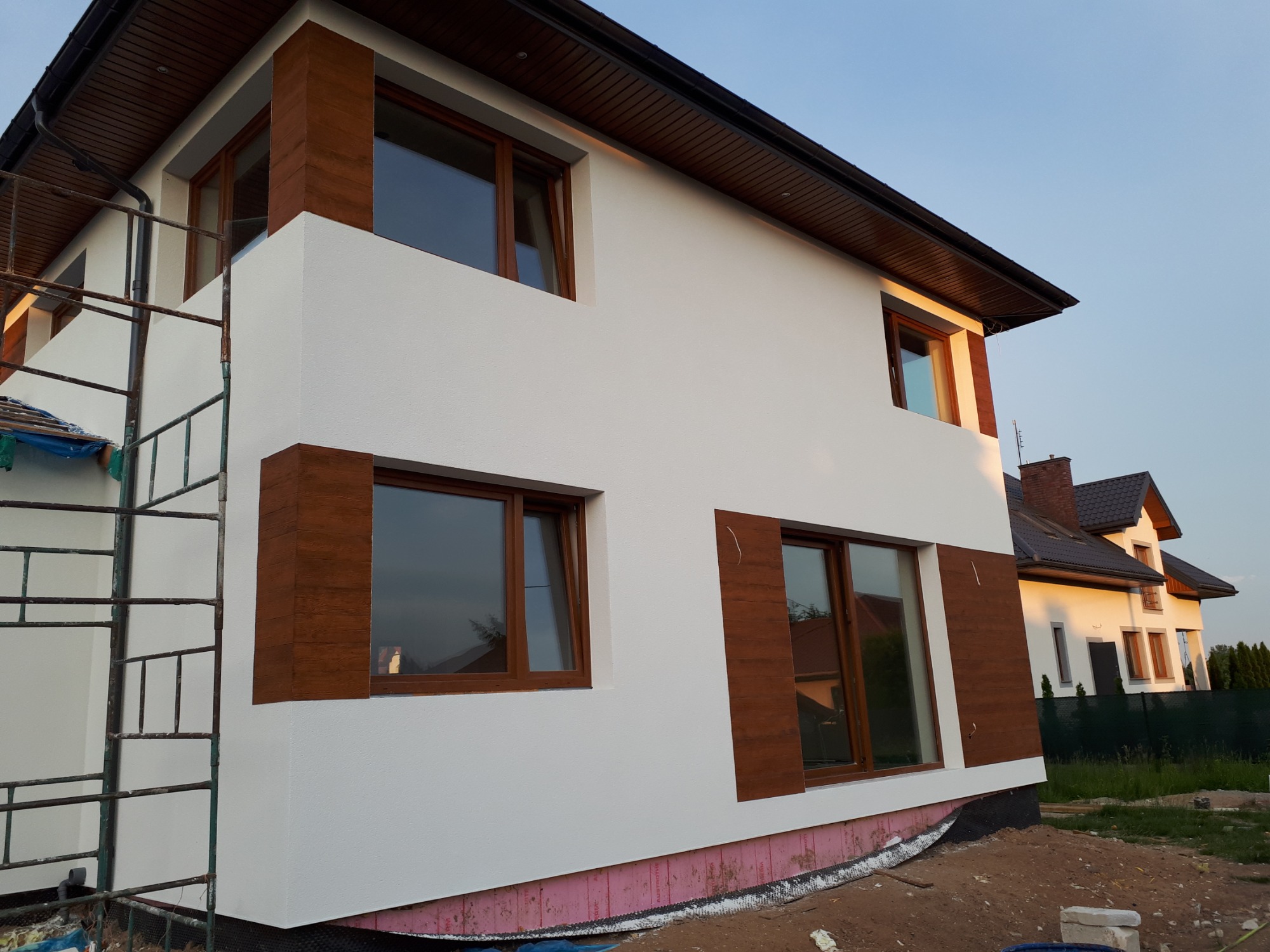 Nowoczesny dom w Warszawie z białymi ścianami i brązowymi elementami dekoracyjnymi wokół okien, widoczne rusztowanie z boku budynku.