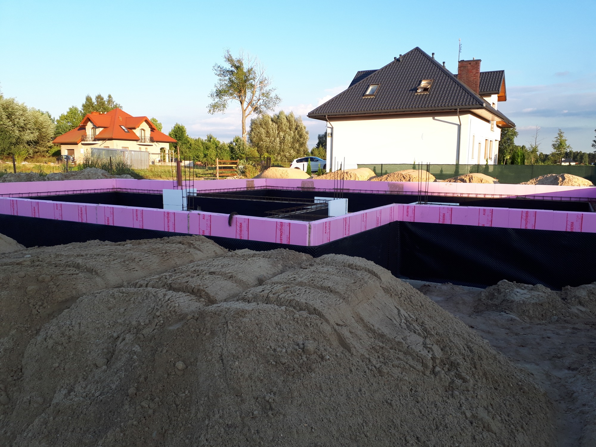Wykop pod fundamenty z widoczną izolacją pionową w postaci czarnej folii i różowych płyt termoizolacyjnych, zbrojenie wewnątrz, w tle domy jednorodzinne w letnim krajobrazie.