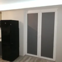 Szare rolety w kasecie zamontowane na białych, dwuskrzydłowych drzwiach balkonowych w nowoczesnej kuchni z czarną lodówką i okapem.