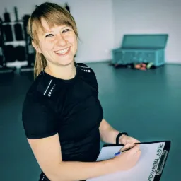 Uśmiechnięta kobieta w sportowym stroju, trzymająca clipboard z napisem 'PLAN TRENINGOWY' i długopisem, w tle sprzęt do ćwiczeń i turkusowa skrzynia.