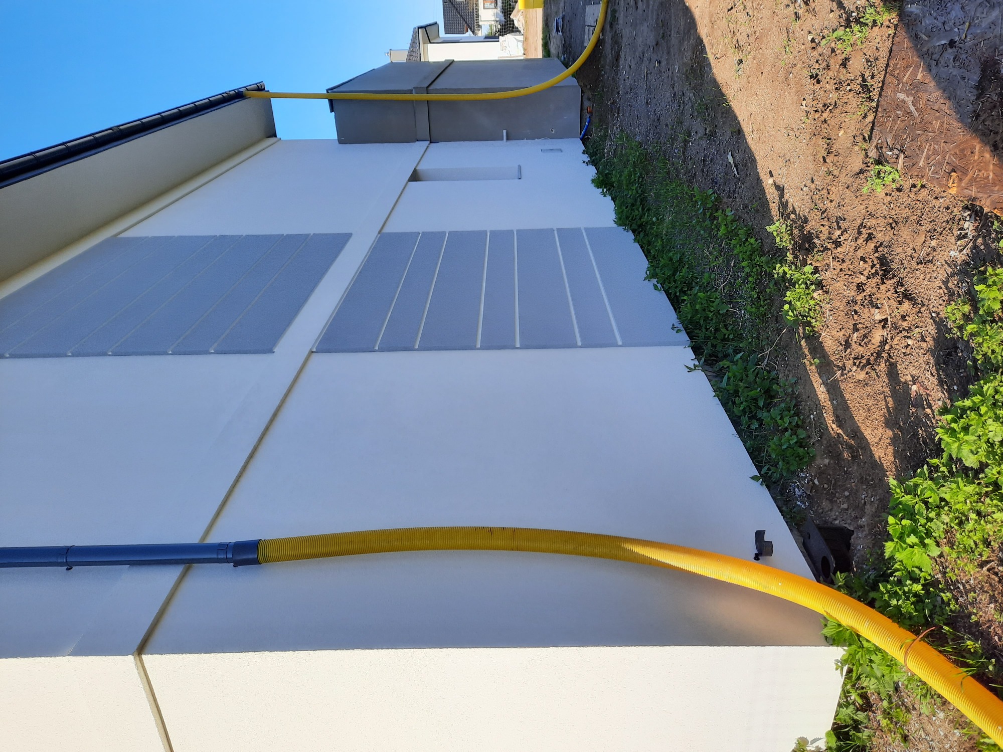 Nowoczesna elewacja budynku z szarymi panelami dekoracyjnymi i żółtym wężem wentylacyjnym, widok z boku w słoneczny dzień.