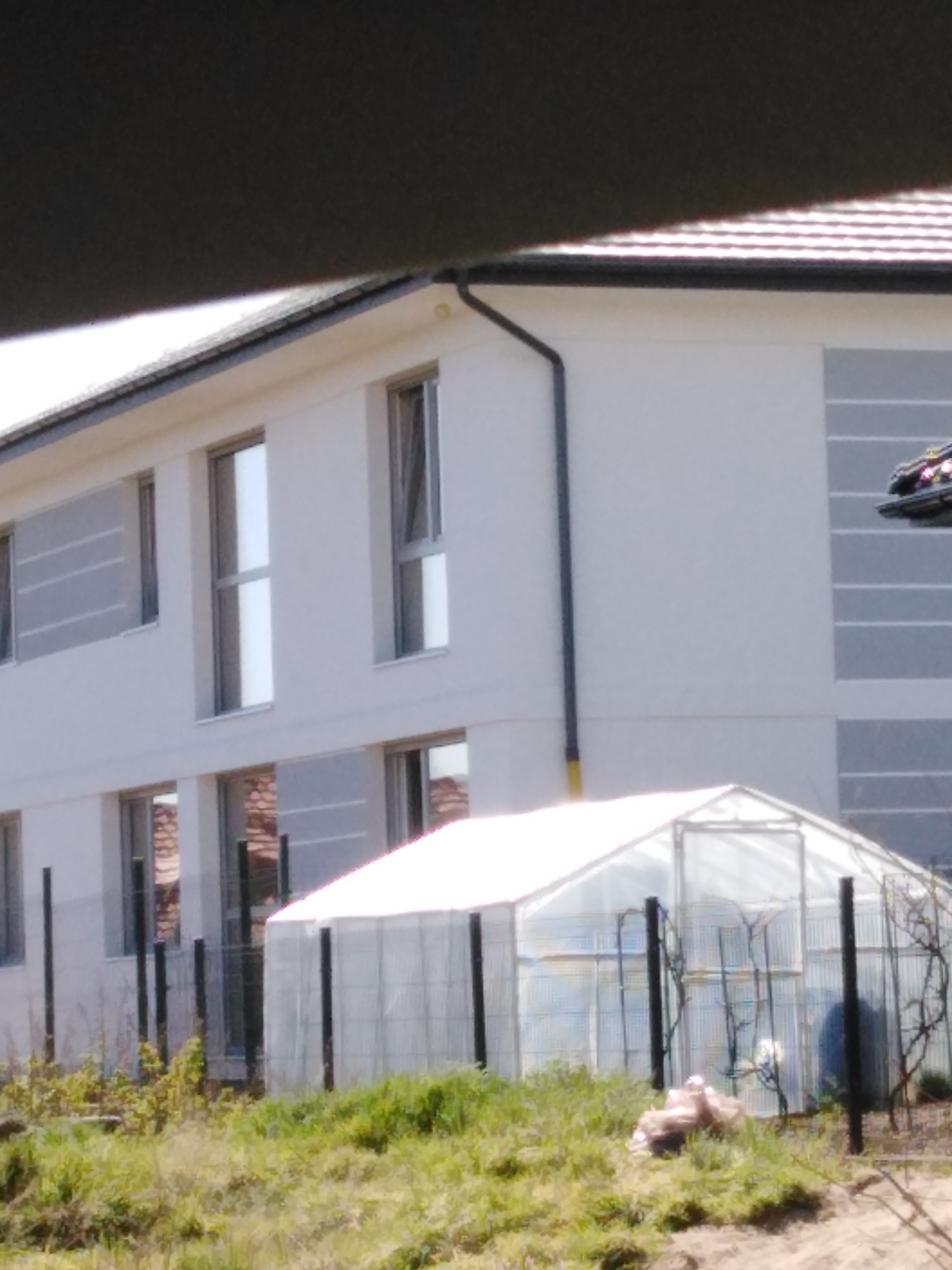 Dwupiętrowy dom z jasną elewacją i szarymi pasami, widoczny fragment dachu i rynny, w tle szklarnia, ujęcie z dołu.