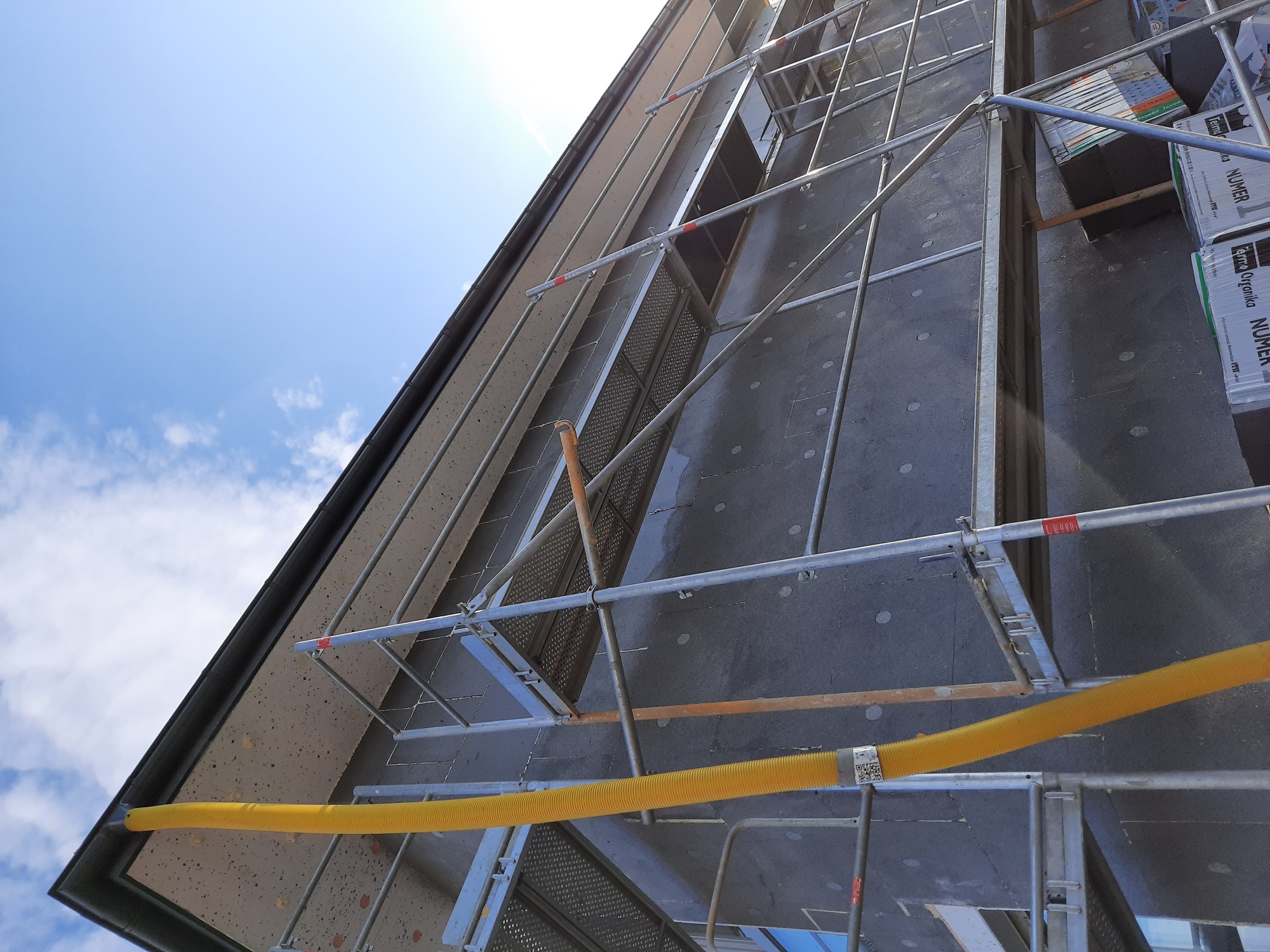 Ocieplanie elewacji budynku z użyciem rusztowania, widoczna żółta rura wentylacyjna i pakiety izolacji termicznej. Ujęcie z dołu, niebo z chmurami w tle.