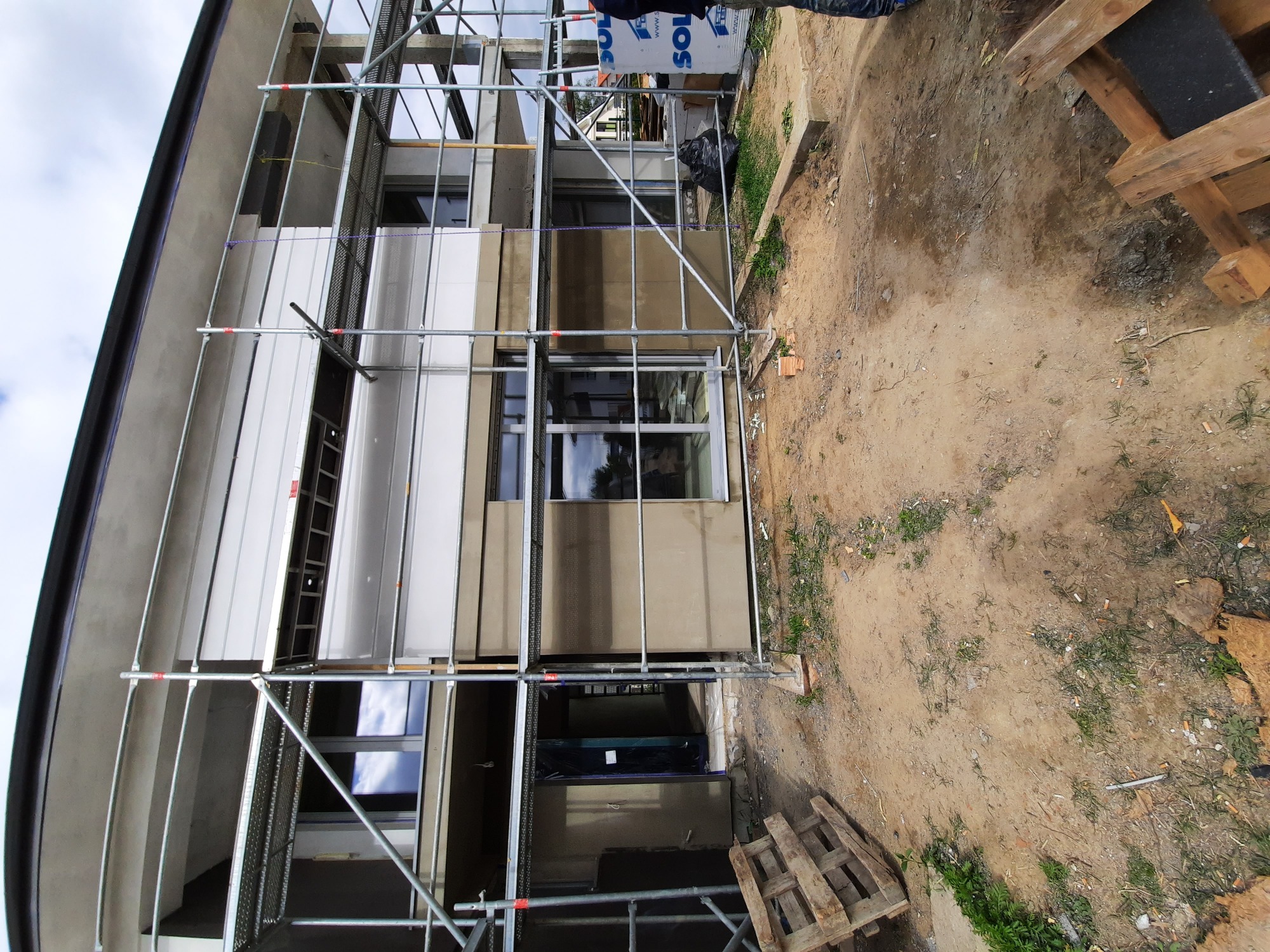Budynek w trakcie prac elewacyjnych, widoczne rusztowania i fragmenty ocieplenia, teren wokół budynku częściowo zagospodarowany.