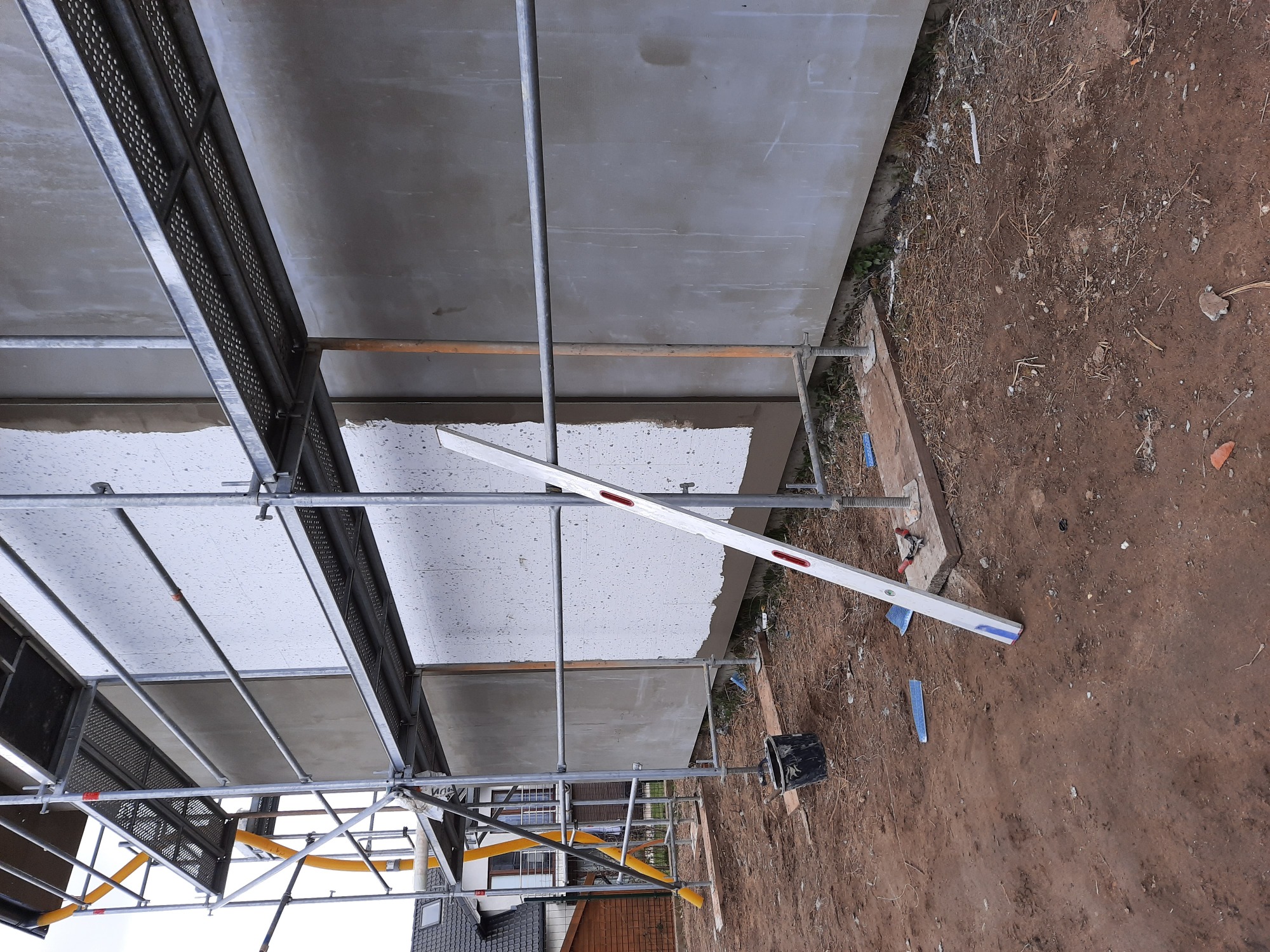 Elewacja budynku w trakcie ocieplania, widoczne rusztowanie, poziomica i grunt obok ściany.