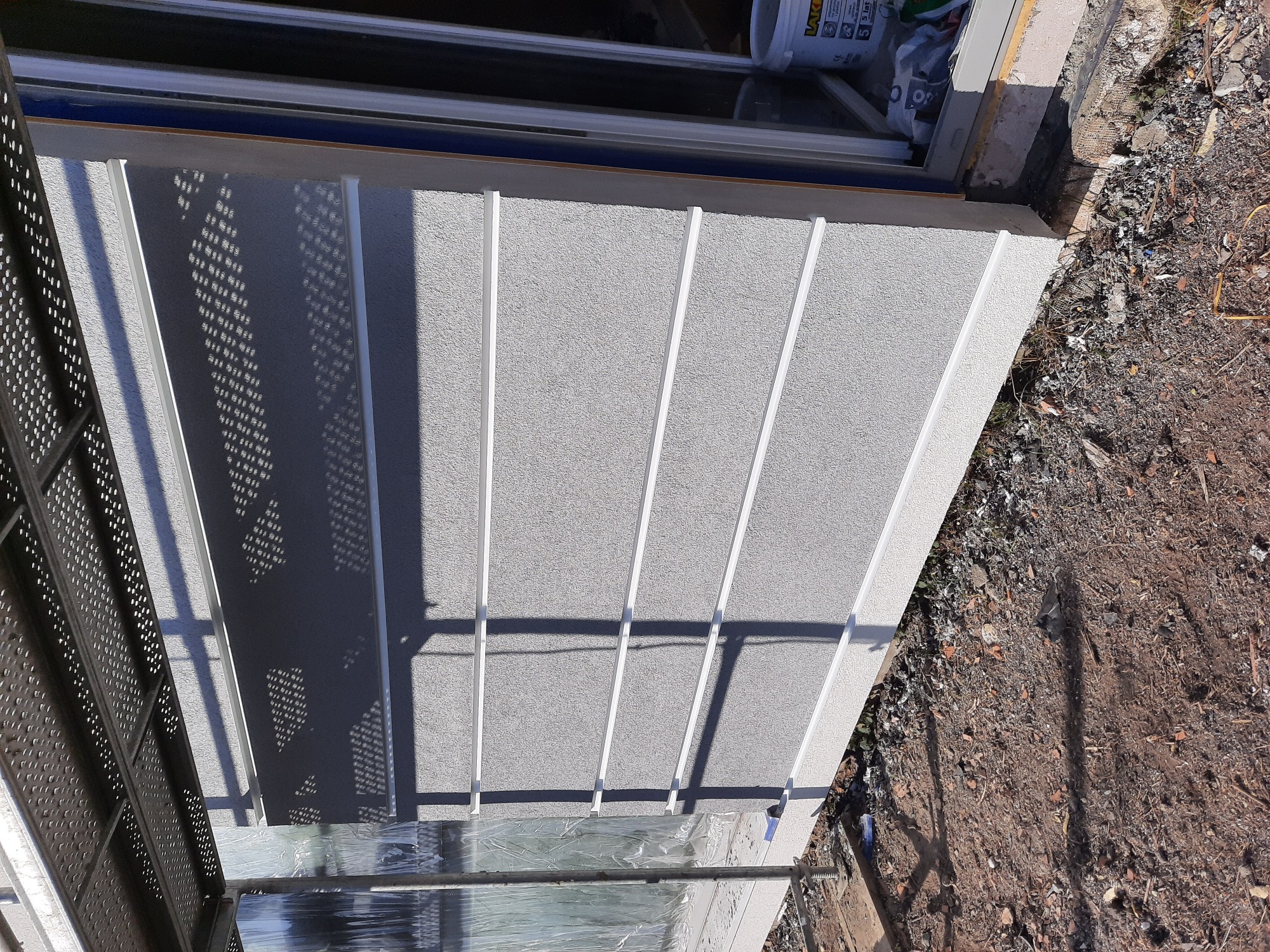 Szara elewacja z wyraźną fakturą i białymi listwami dekoracyjnymi, częściowo zacieniona przez metalową konstrukcję rusztowania. Widoczne wiadro z farbą i gruz przy ścianie.