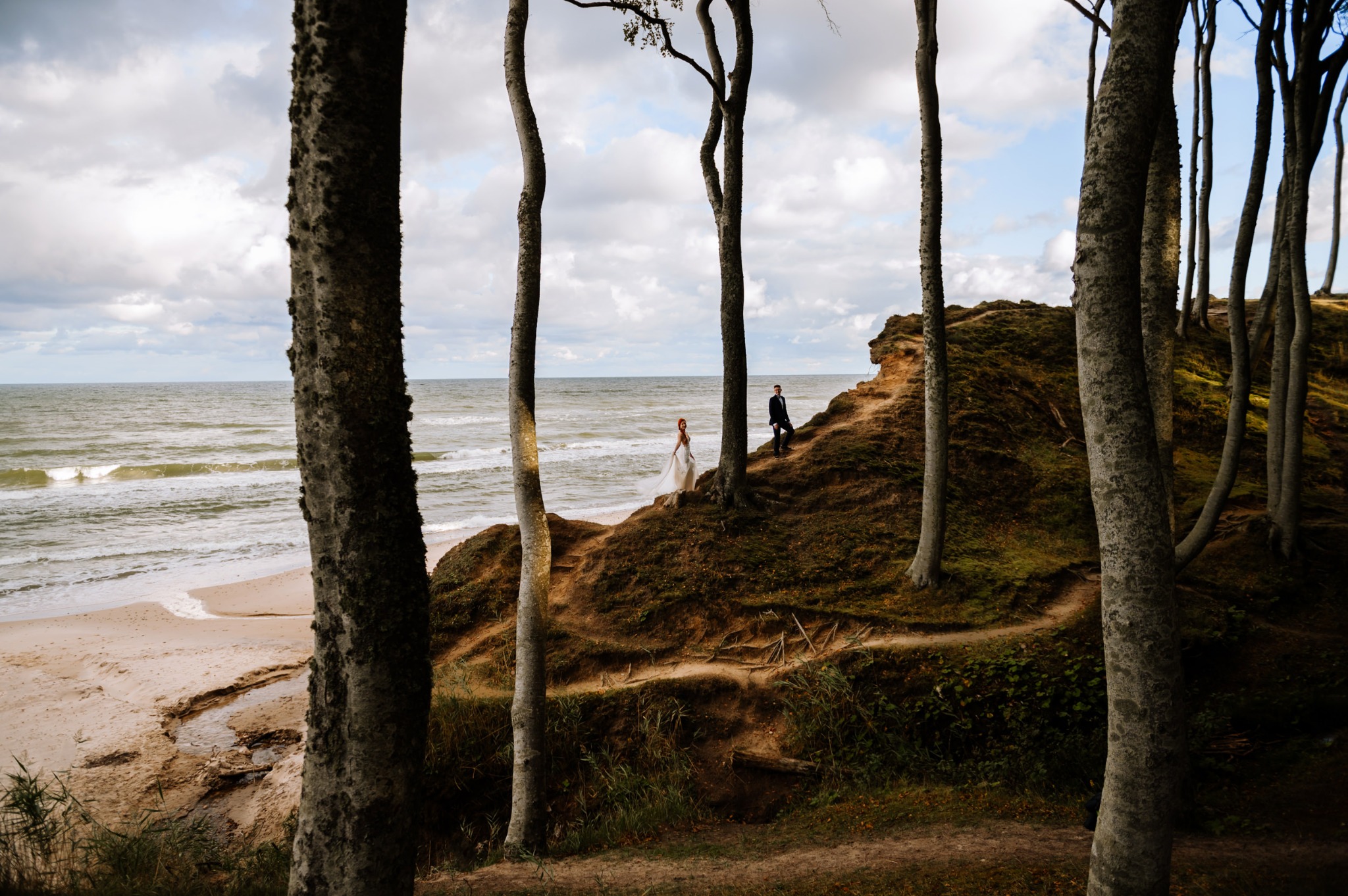 Para młoda w plenerze: panna młoda w zwiewnej sukni i pan młody w garniturze spacerują po klifowym brzegu morza, widoczni pomiędzy drzewami, z plażą i falami w tle.