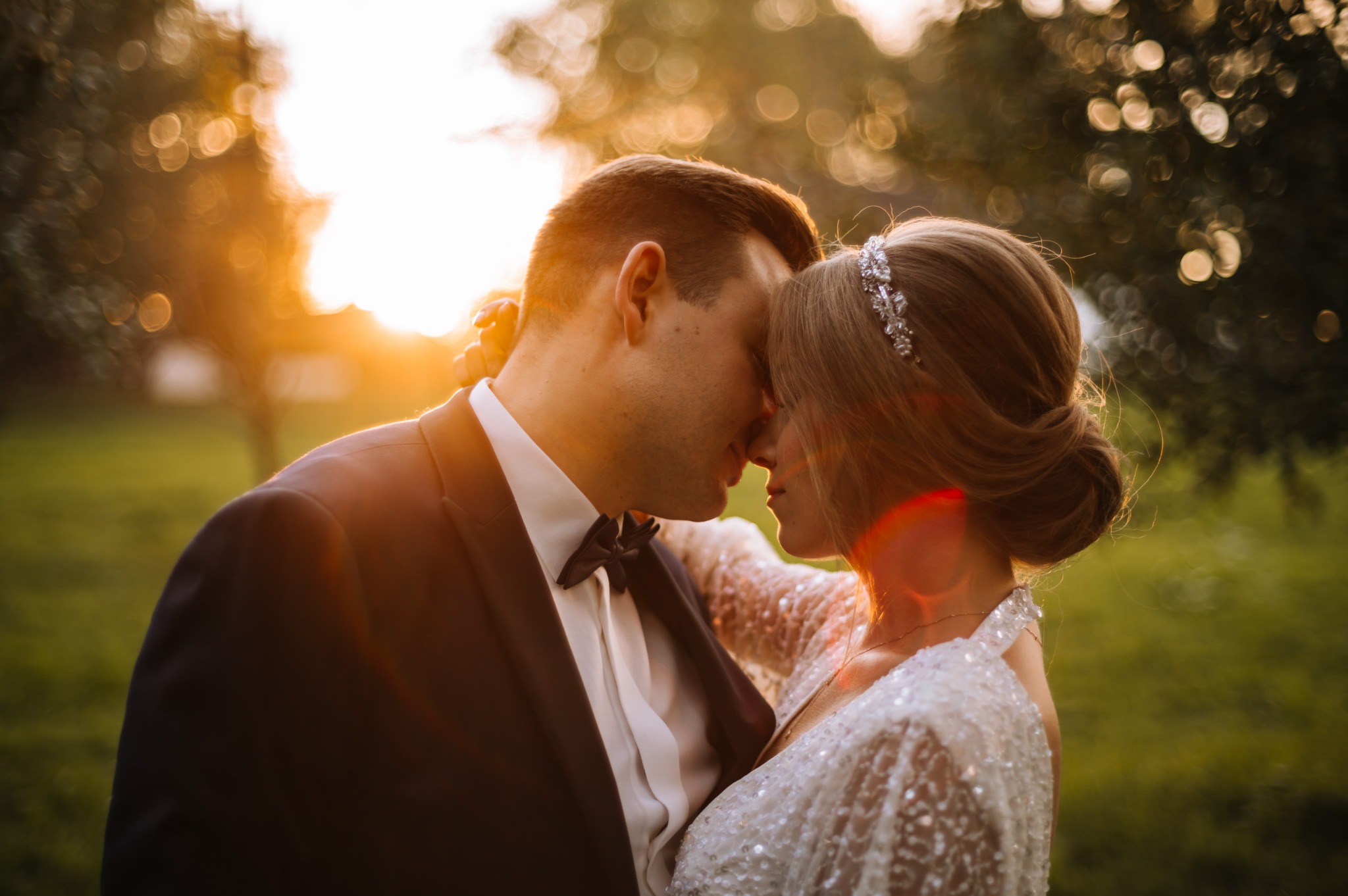 Para młoda w objęciach, zbliżenie twarzy, on w czarnym garniturze z muchą, ona w białej sukni z cekinami i ozdobą we włosach, ciepłe światło zachodzącego słońca.