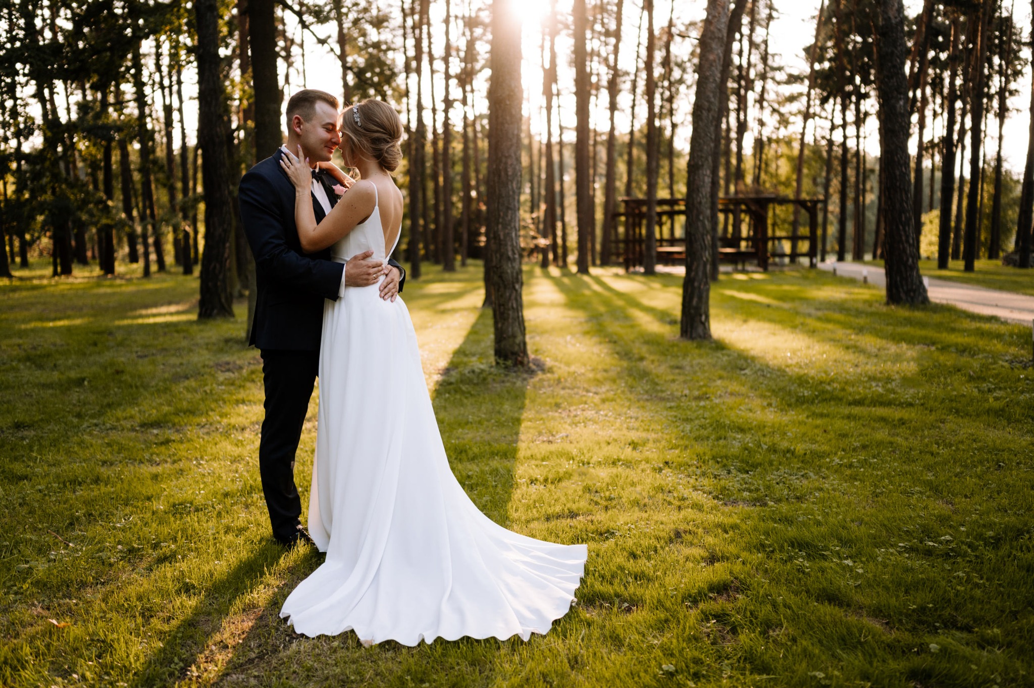 Para młoda w objęciach na tle leśnej scenerii, promienie słońca przebijają przez drzewa, ona w białej sukni z odkrytymi plecami, on w czarnym garniturze.