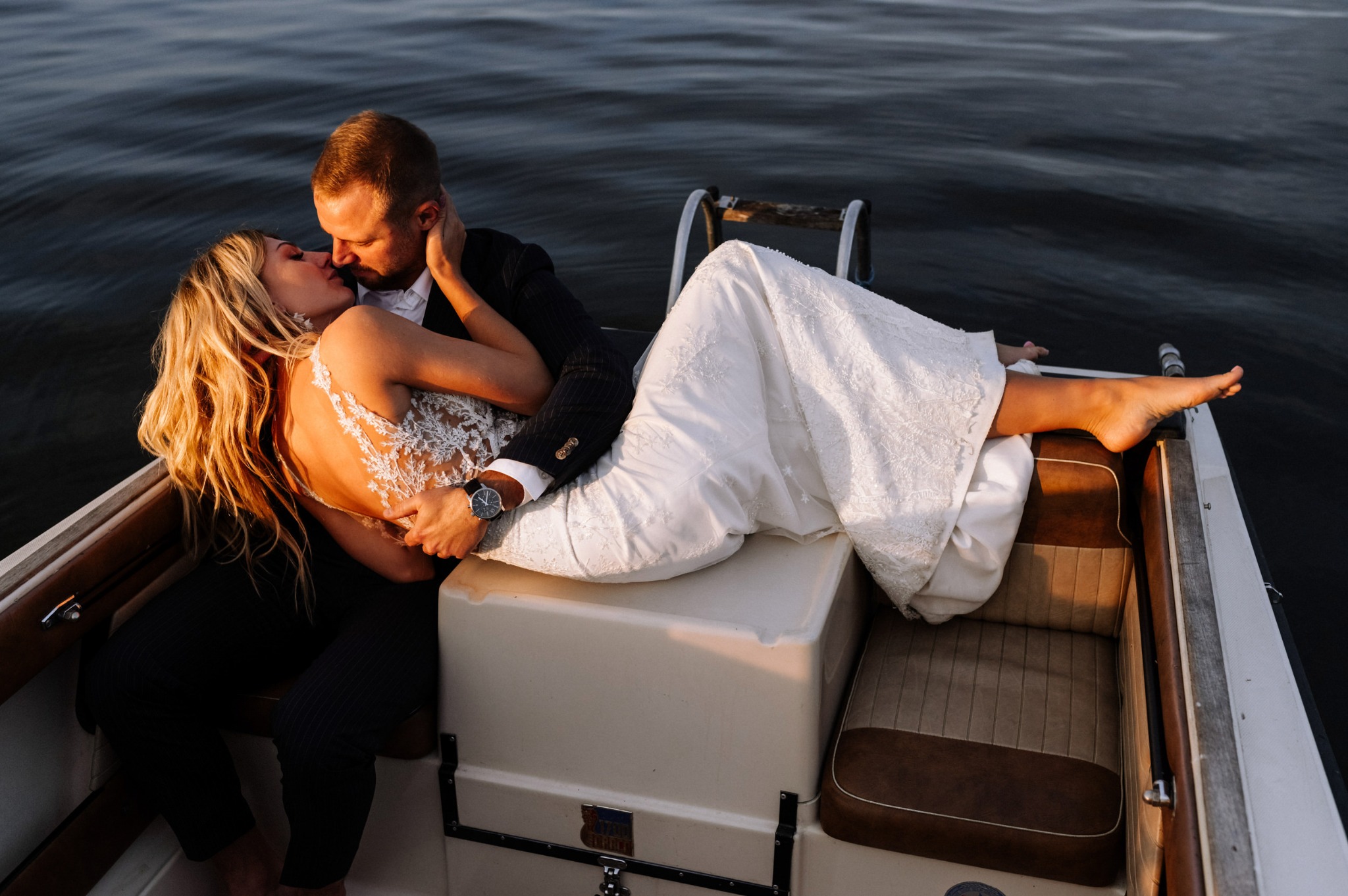 Para młoda w łódce podczas pocałunku; suknia ślubna z koronkowymi plecami, garnitur w prążki, zachód słońca nad wodą.