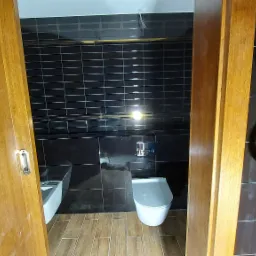 Wykończona łazienka z czarnymi, prostokątnymi płytkami na ścianach, drewnopodobną podłogą i białymi sanitariatami, widziana przez otwarte drewniane drzwi.