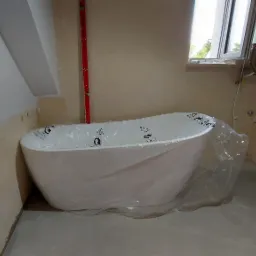 Nowa biała wanna owalna, zabezpieczona folią ochronną, w surowym wnętrzu łazienki w trakcie prac wykończeniowych, widoczna instalacja grzewcza i płyta kartonowo-gipsowa z instalacją hydrauliczną.