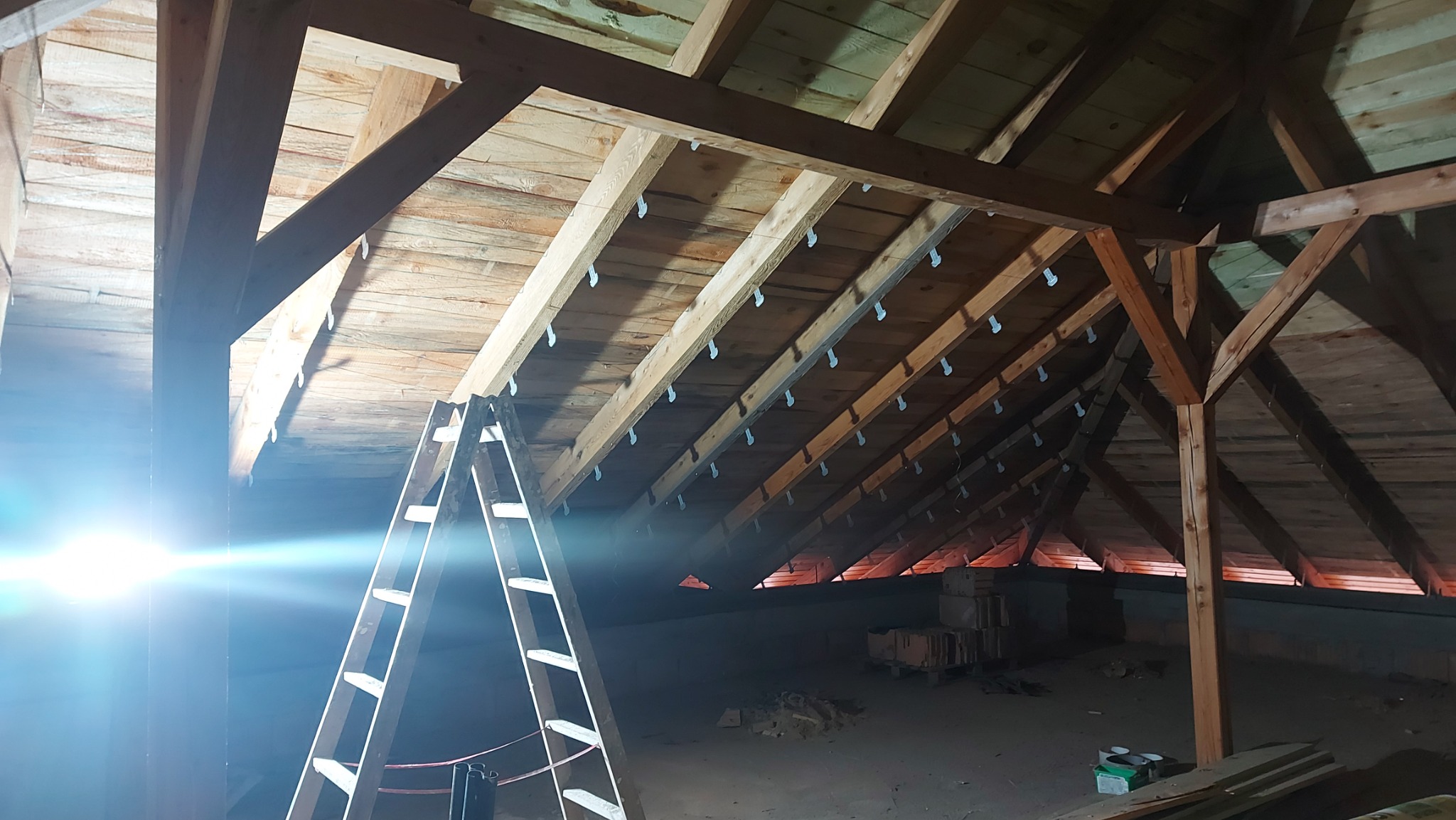 Surowe drewniane belki stropowe na poddaszu z widocznymi metalowymi mocowaniami, drabina oparta o konstrukcję, przygotowane materiały budowlane w tle.