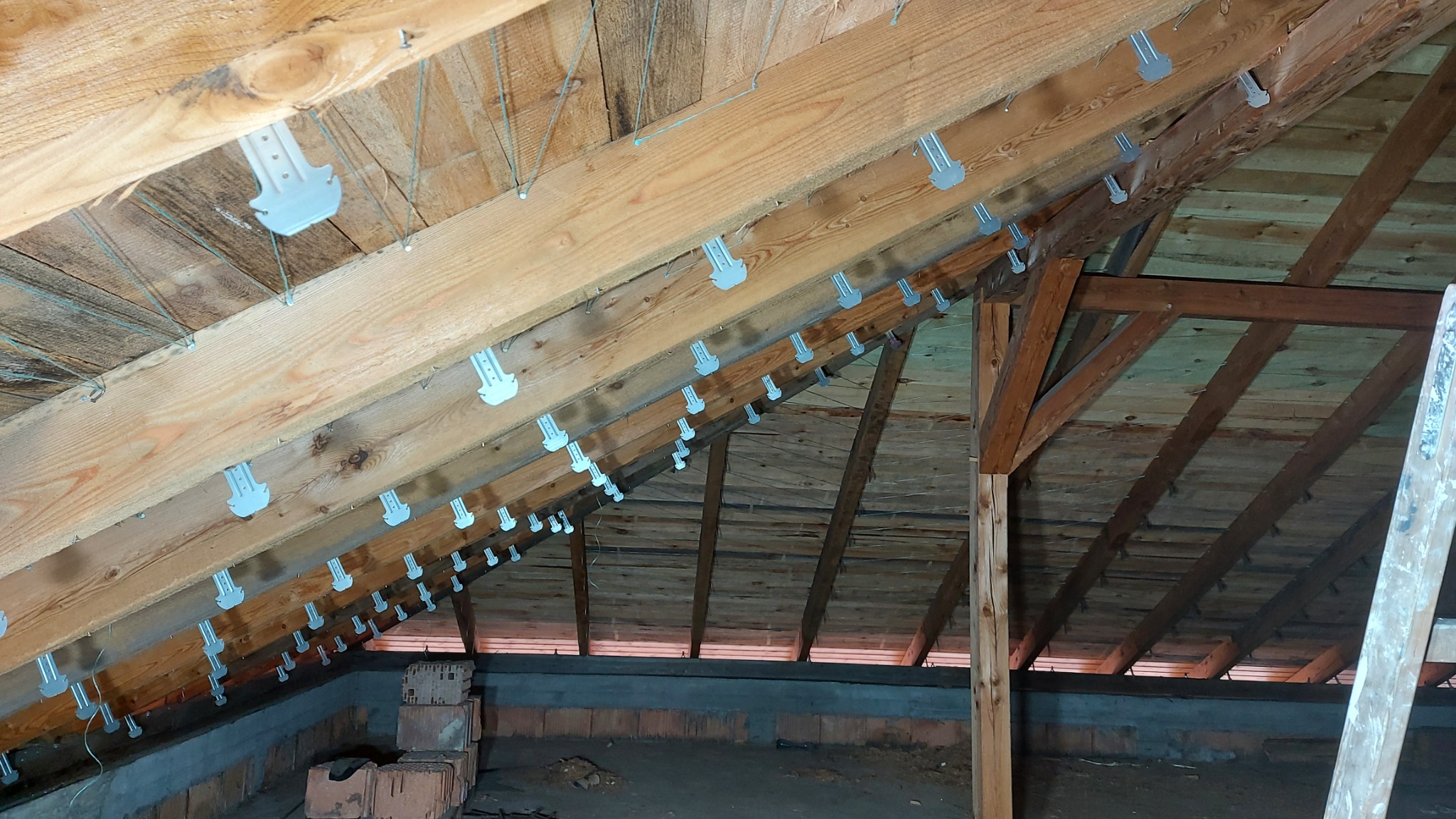 Konstrukcja drewniana poddasza z zamocowanymi uchwytami do montażu płyt gipsowo-kartonowych, widoczne cegły i bloczki betonowe w dolnej części.