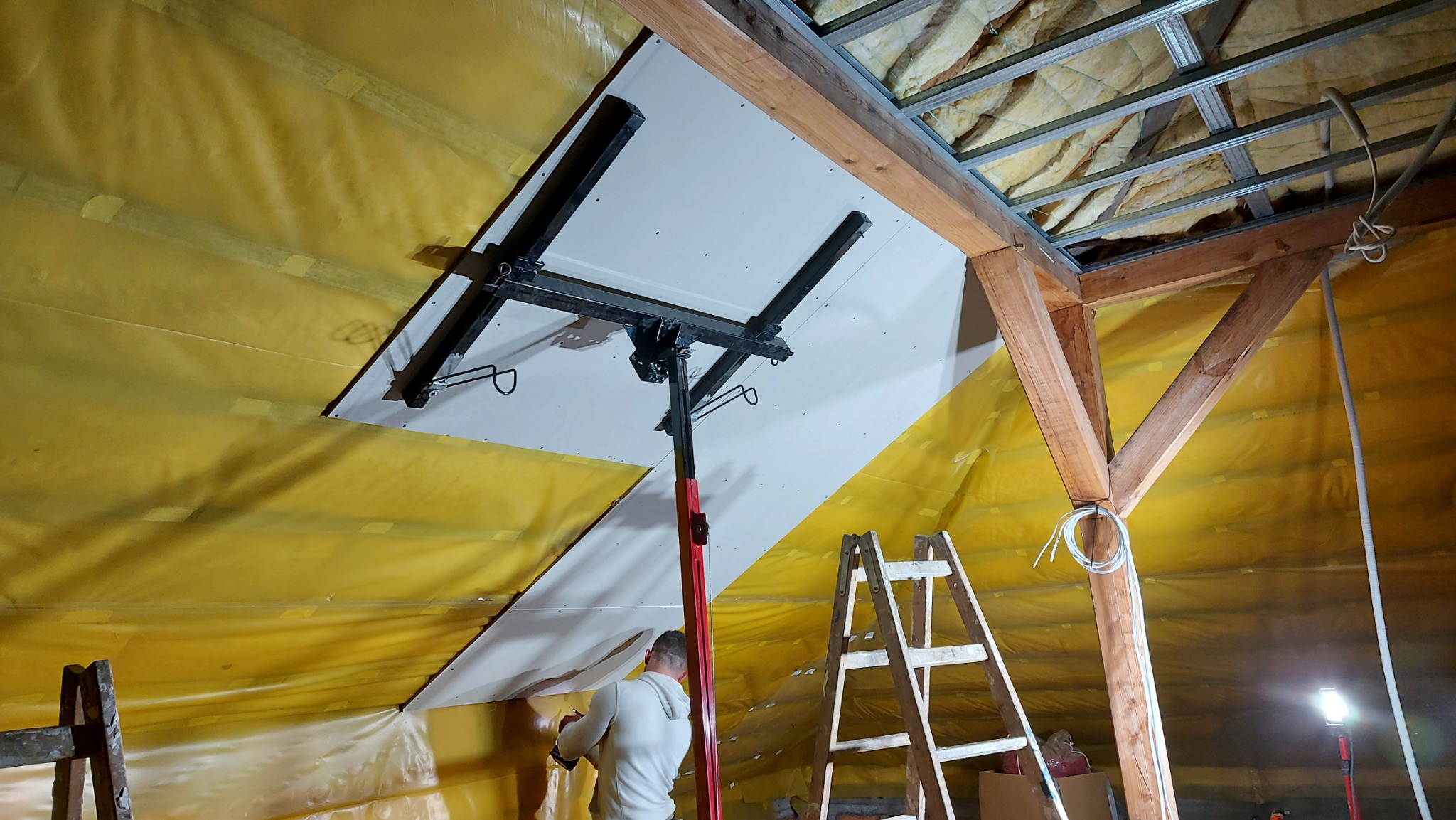 Montaż płyty gipsowo-kartonowej na skośnym suficie poddasza z użyciem windy do płyt, widoczna izolacja z folii paroizolacyjnej, drewniana konstrukcja dachu oraz drabina.