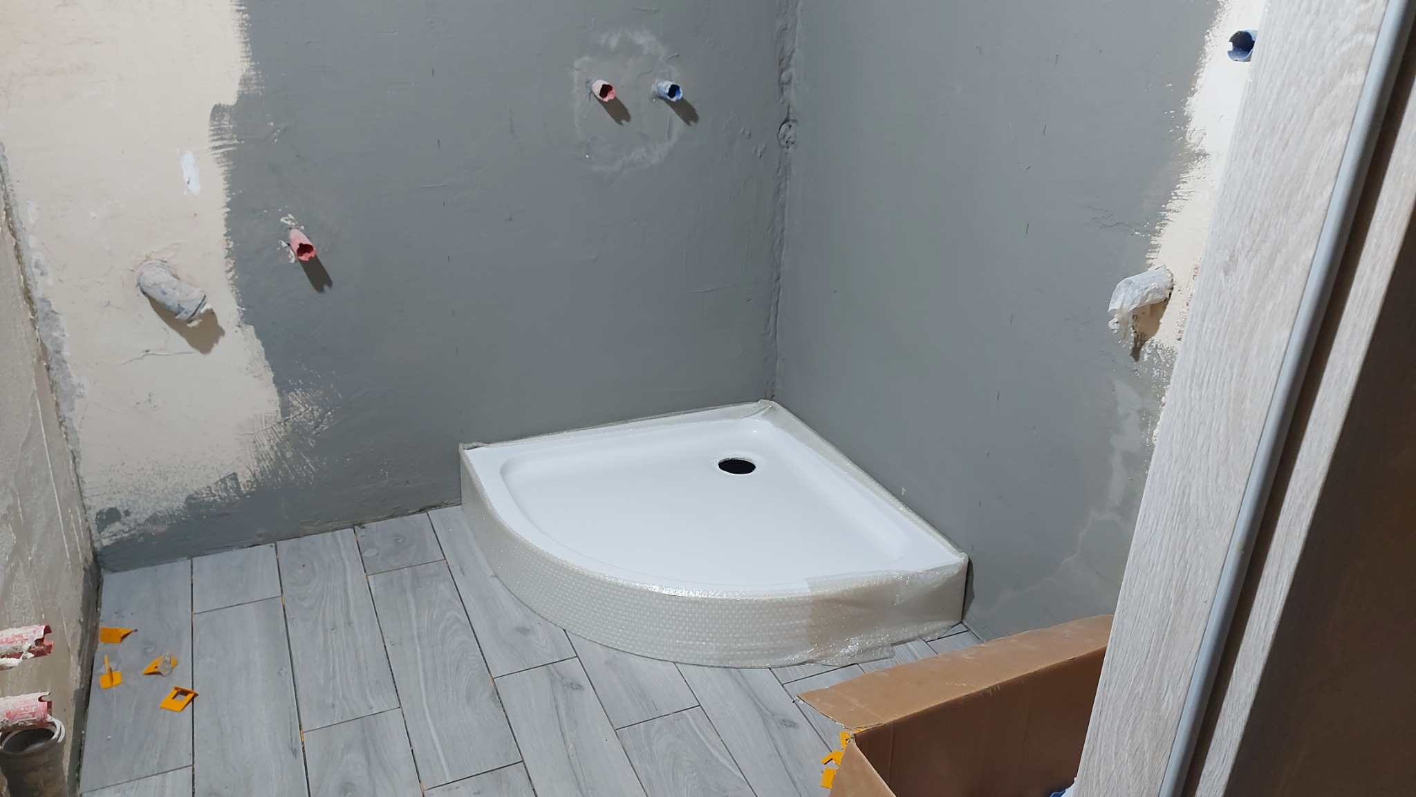 Narożny brodzik owinięty folią ochronną w surowej łazience w trakcie remontu, widoczne przygotowane ściany z instalacjami, szare płytki podłogowe imitujące drewno oraz elementy kartonu.