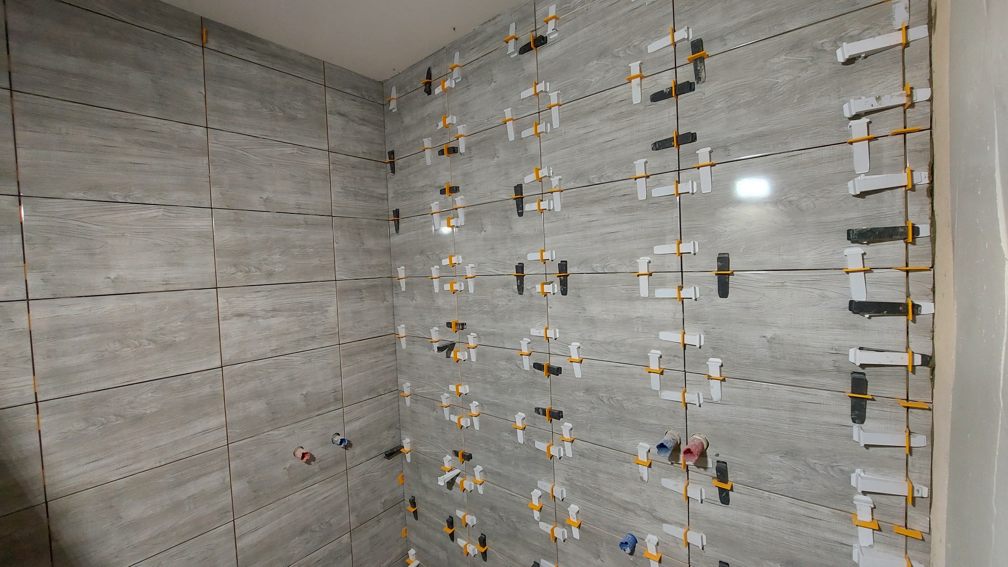 Ściana łazienki w trakcie kładzenia płytek imitujących drewno, z widocznymi krzyżykami dystansowymi i poziomującymi, przyłącza wodne wystają ze ściany.