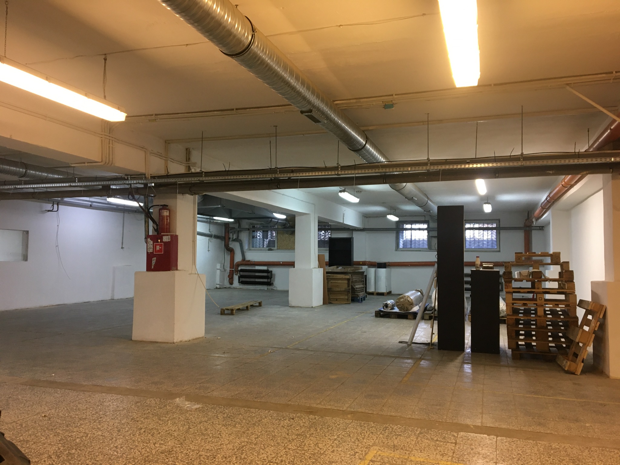 Przestronne, puste pomieszczenie magazynowe z widocznymi elementami instalacji pod sufitem, w tym rurami wentylacyjnymi i przewodami elektrycznymi. Na ścianie widoczna skrzynka przeciwpożarowa.