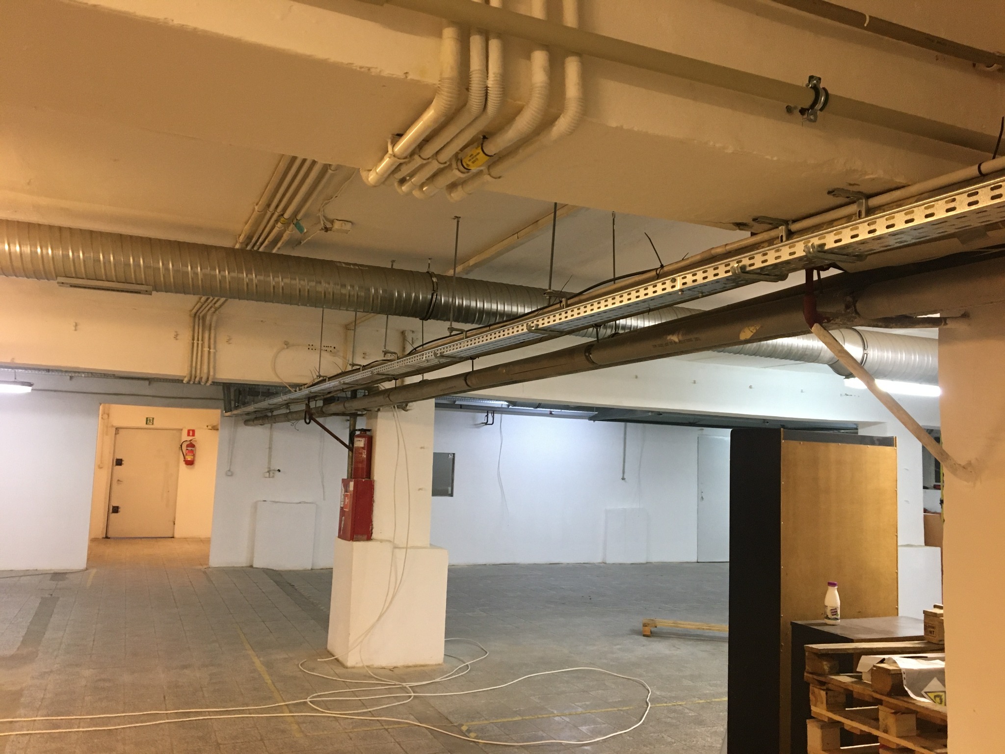 Metalowe i plastikowe rury wentylacyjne oraz koryta kablowe pod sufitem w hali przemysłowej, widoczny także hydrant i gaśnica.