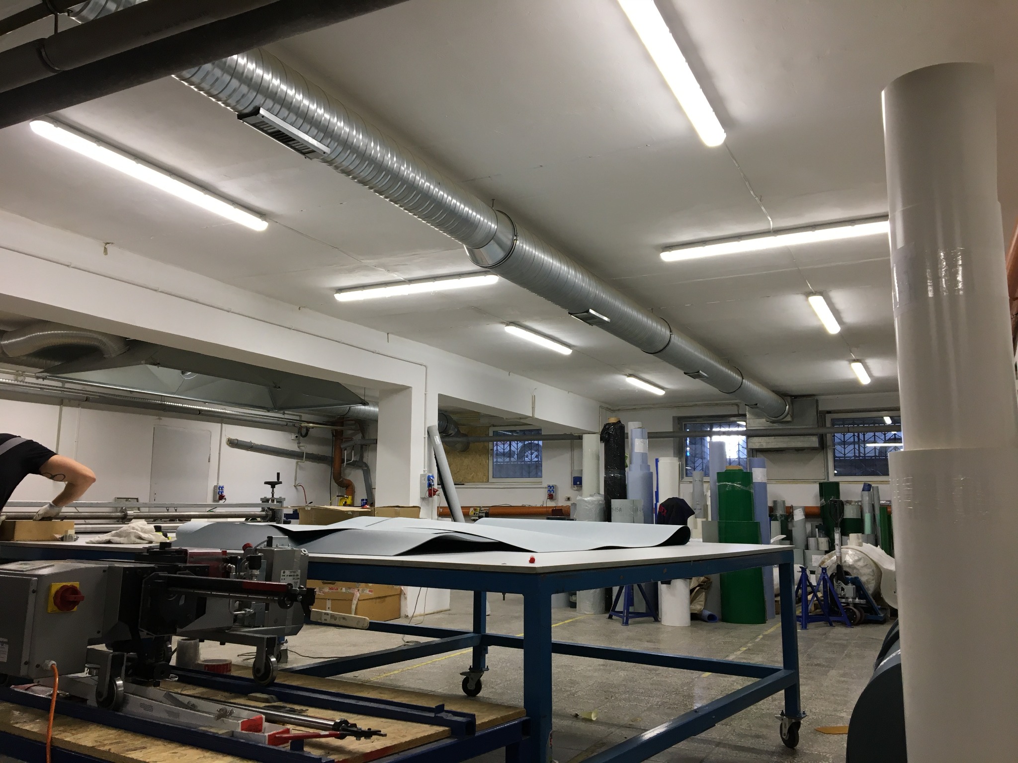 Warsztat z maszyną do cięcia i stołem z szarym materiałem, widoczne rury wentylacyjne pod sufitem i okna w tle.