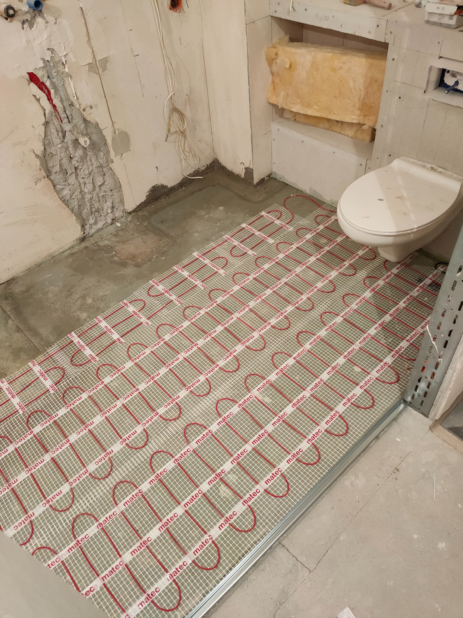 Montaż maty grzewczej Matec z czerwonymi przewodami na siatce, widok na surową łazienkę w trakcie remontu, z widocznymi przewodami elektrycznymi i fragmentem toalety.
