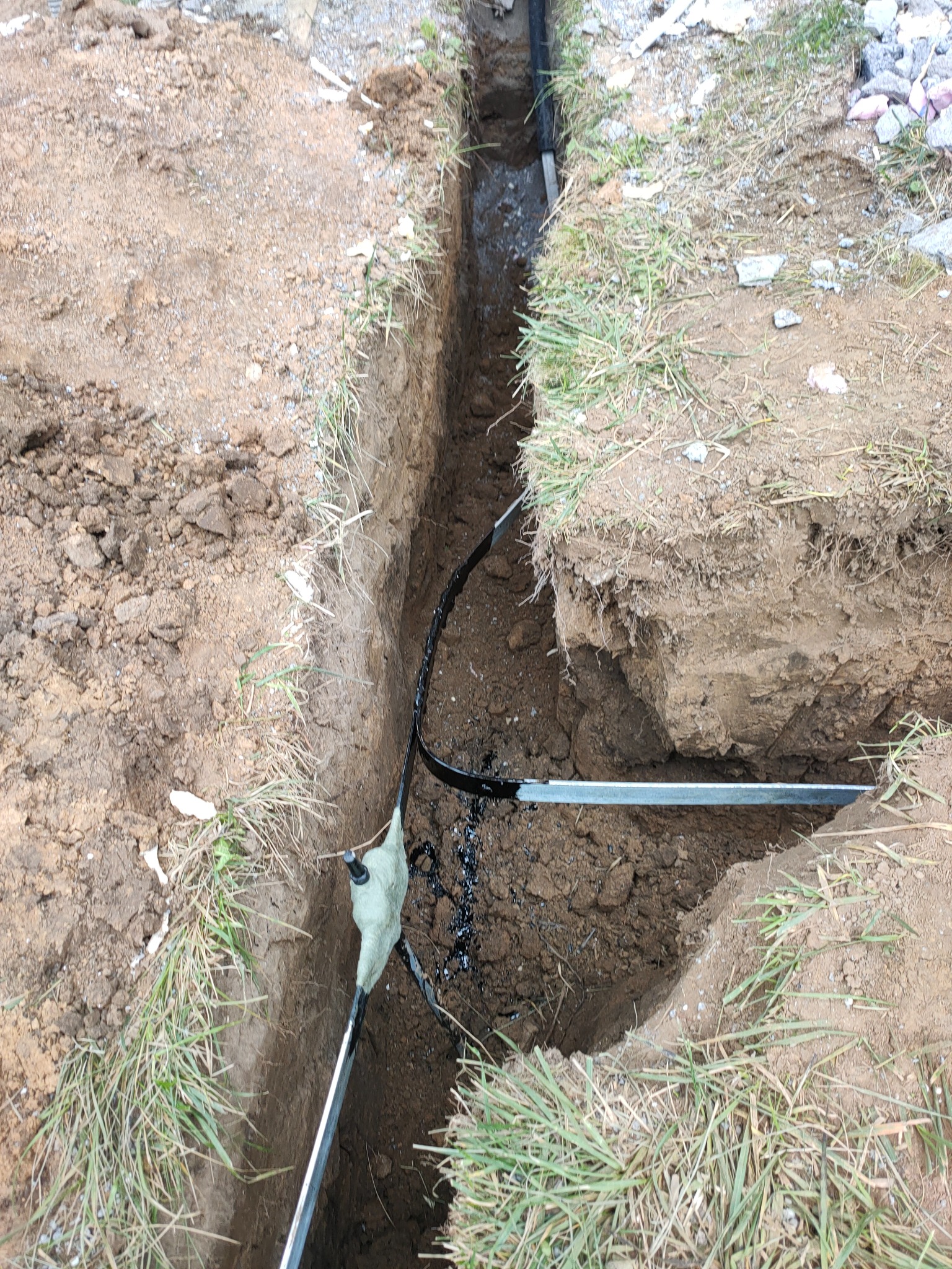 Wkopany w ziemię kabel elektryczny w wykopie, zabezpieczony izolacją, widoczne narzędzia i ziemia.