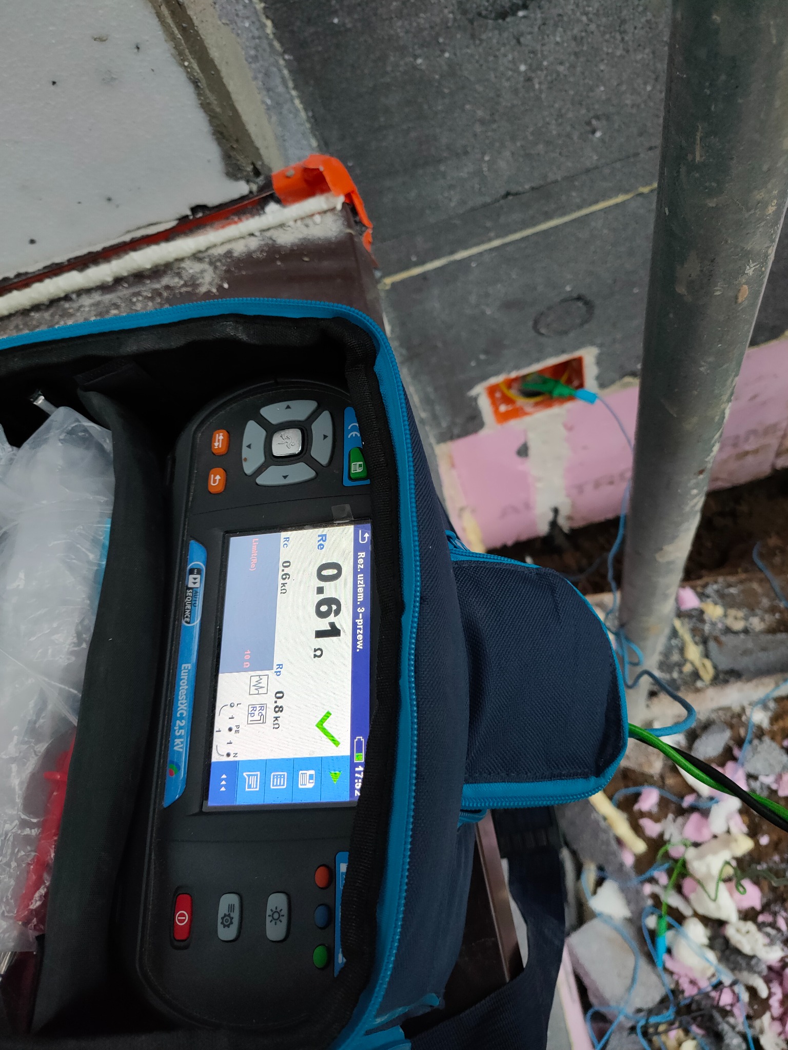 Miernik EurotestXC 2.5 kV w torbie pomiarowej, wyświetlający wynik 0.61 Ω, z podłączonymi przewodami do puszki elektrycznej w ścianie ocieplonej styropianem, na tle rusztowania.