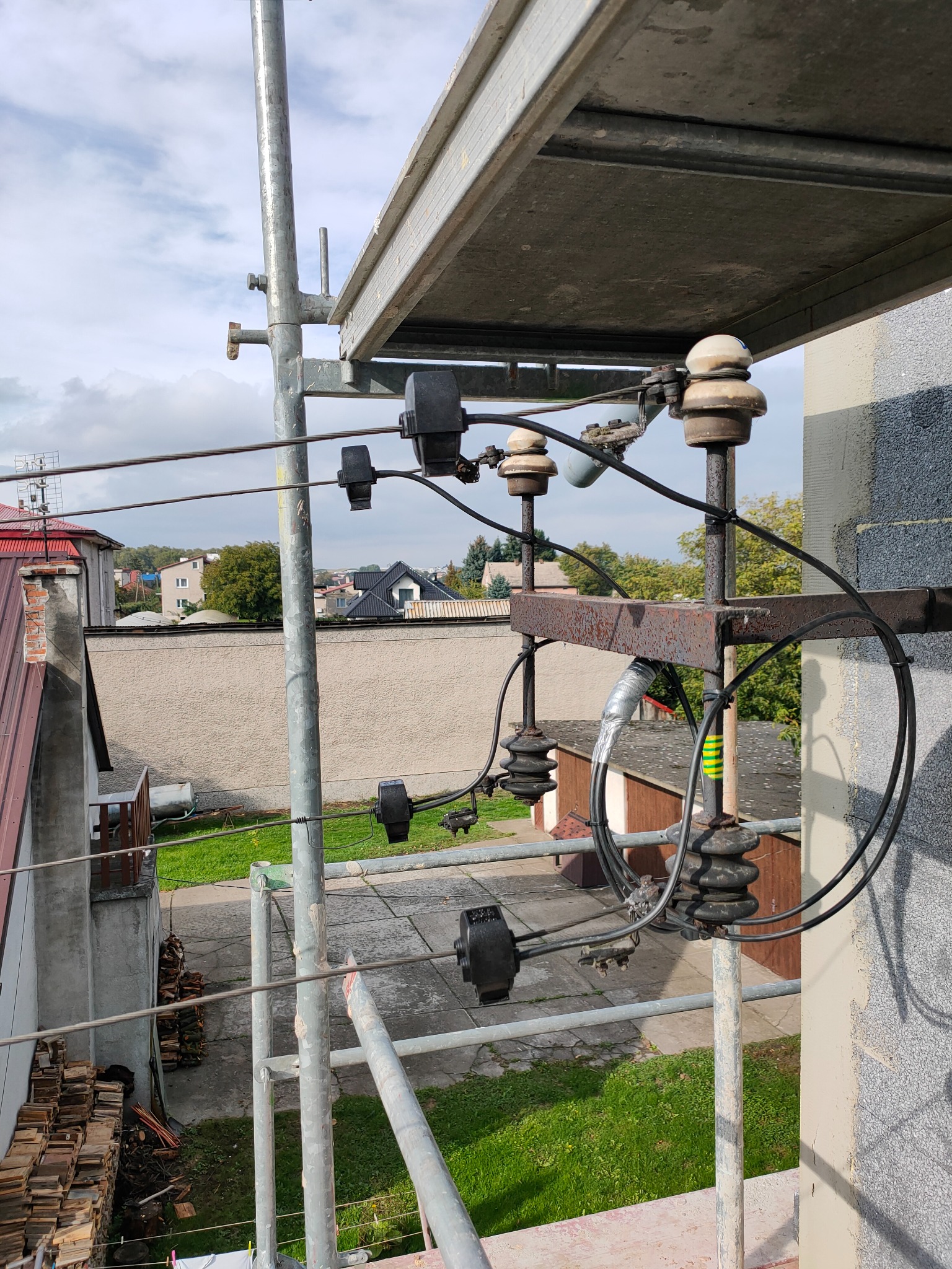 Zestaw izolatorów i przewodów elektrycznych zamontowanych na metalowej konstrukcji, widoczny z rusztowania na tle zabudowań i zieleni.
