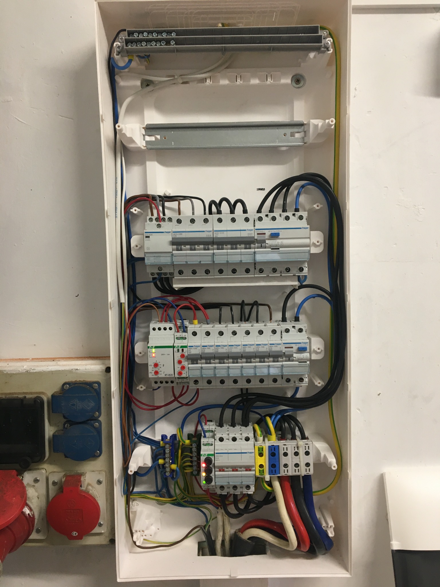 Otwarta skrzynka rozdzielcza z zamontowanymi bezpiecznikami i przewodami o różnych kolorach, widoczne także gniazda siłowe na ścianie obok.