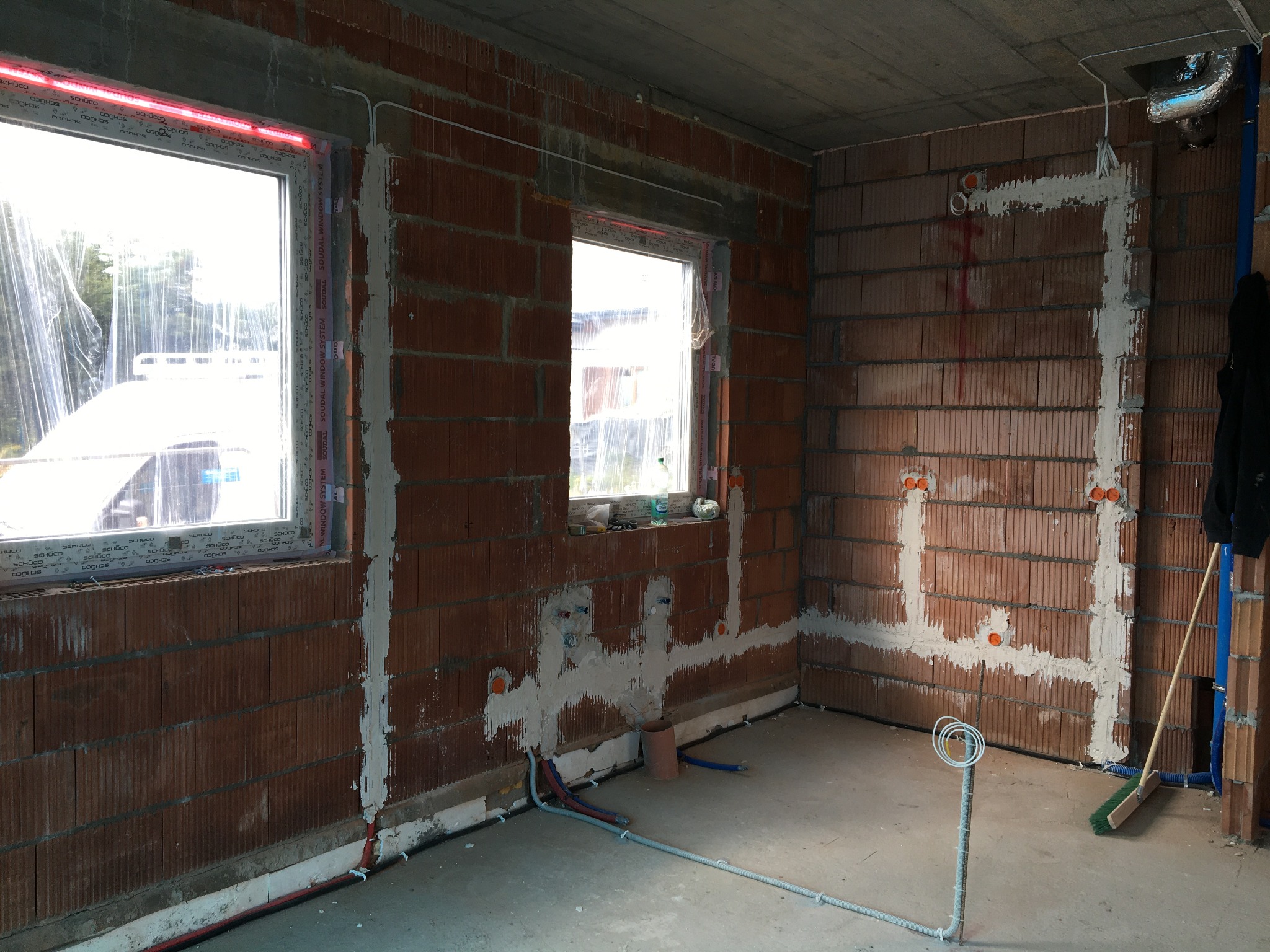 Wnętrze w trakcie rozprowadzania przewodów elektrycznych w nowym budynku z cegły, widoczne puszki instalacyjne i kable prowadzone po ścianach, okna zabezpieczone folią.