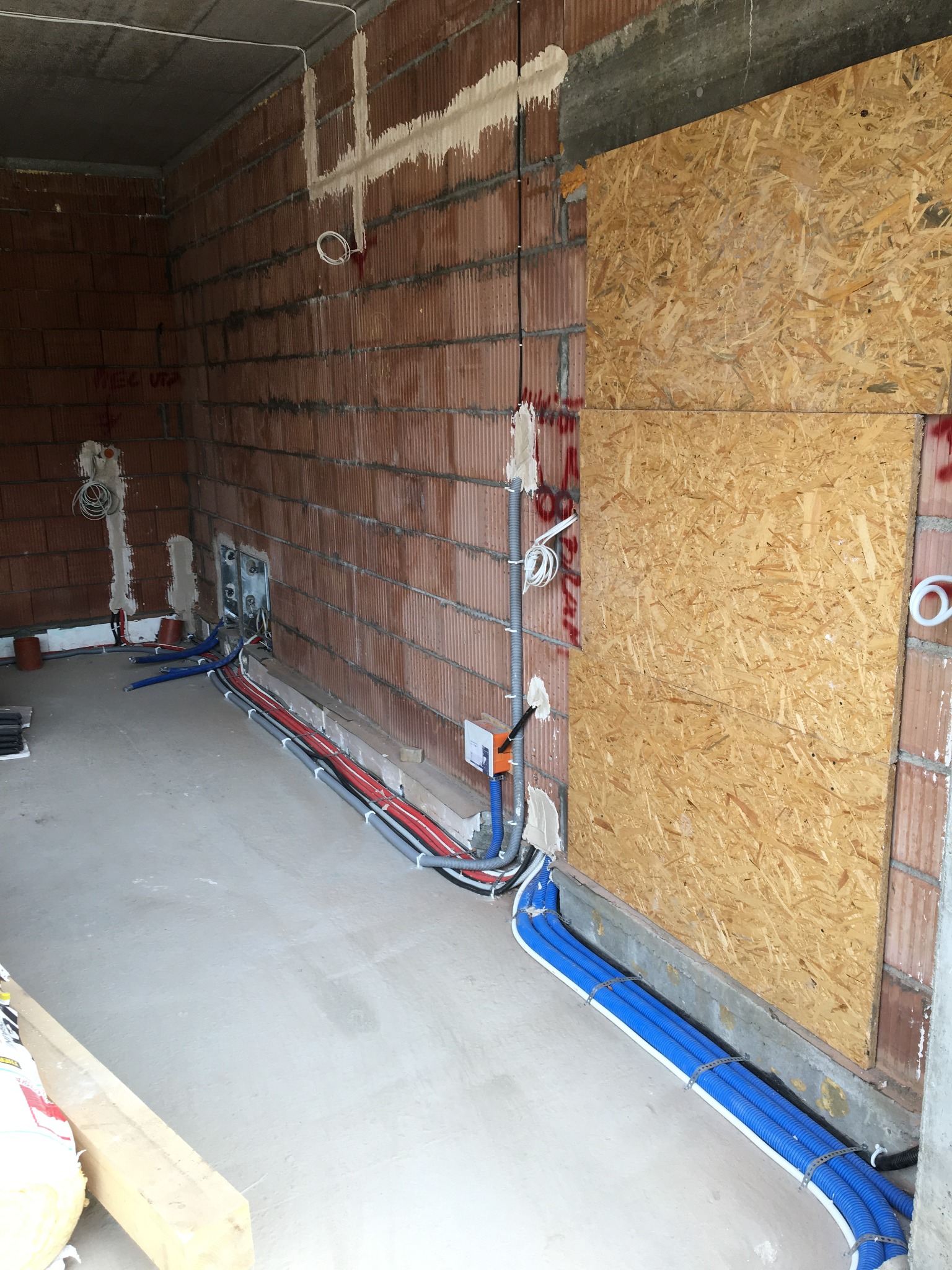 Wnętrze w trakcie instalacji elektrycznej: kable w peszlach (niebieskie, czerwone, szare) ułożone wzdłuż ściany z cegły dziurawki, zabezpieczone mocowaniami; widoczne puszki instalacyjne i fragment...