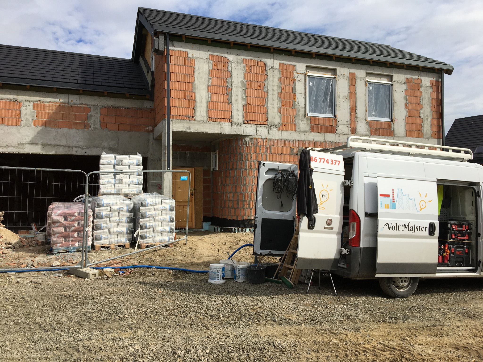 Budynek w trakcie budowy z widocznym ociepleniem muru, obok zaparkowany biały van z logo firmy 'Volt Majster' i numerem telefonu, otwarte tylne drzwi odsłaniają narzędzia wewnątrz.