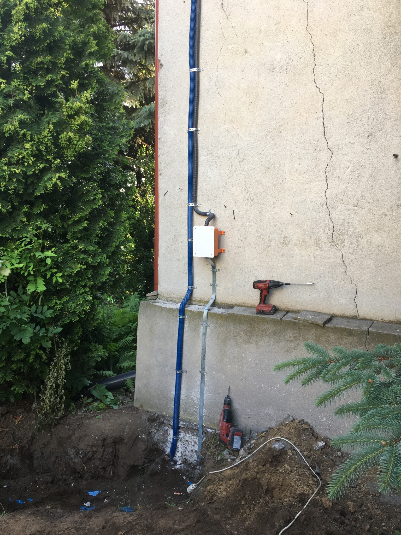 Niebieskie kable w karbowanej osłonie przymocowane do zewnętrznej ściany budynku, z puszką przyłączeniową i narzędziami elektrycznymi leżącymi na ziemi w trakcie prac.
