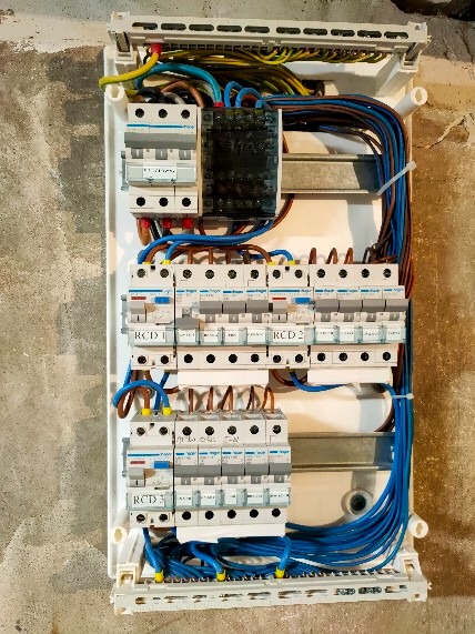 Otwarta skrzynka elektryczna z zainstalowanymi bezpiecznikami różnicowoprądowymi (RCD 1, RCD 2, RCD 3) i licznymi przewodami w kolorach: niebieskim, brązowym i żółto-zielonym, zamocowana na szarej,...