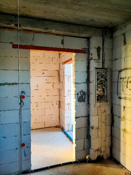 Surowy stan budynku w trakcie prac wykończeniowych, widoczna puszka elektryczna z przewodami na ścianie z białego bloczka, przewody prowadzone wzdłuż ściany, zarys przejścia do innego pomieszczenia.