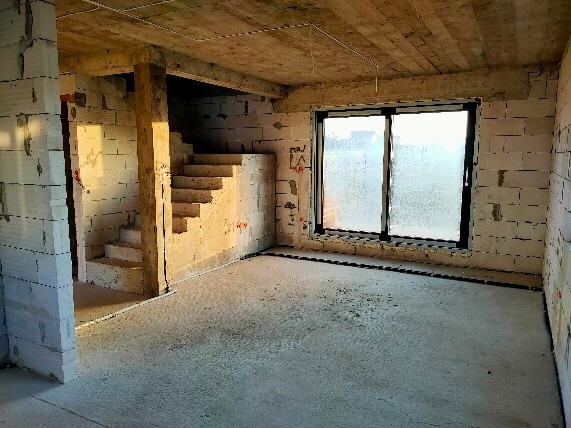 Surowy stan wnętrza domu w trakcie budowy: ściany z białego bloczka, betonowa podłoga i schody, widoczne przewody na suficie, okno z zaparowaną szybą.