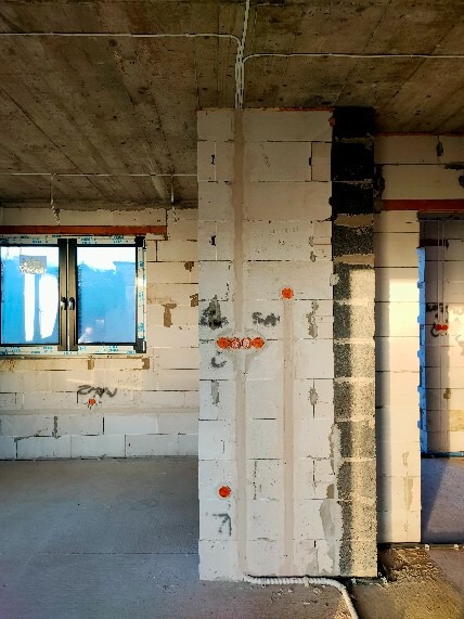 Surowy stan wnętrza budynku z widocznymi białymi ścianami z betonu komórkowego, oznaczonymi miejscami pod gniazdka elektryczne, oraz kable prowadzone po suficie.