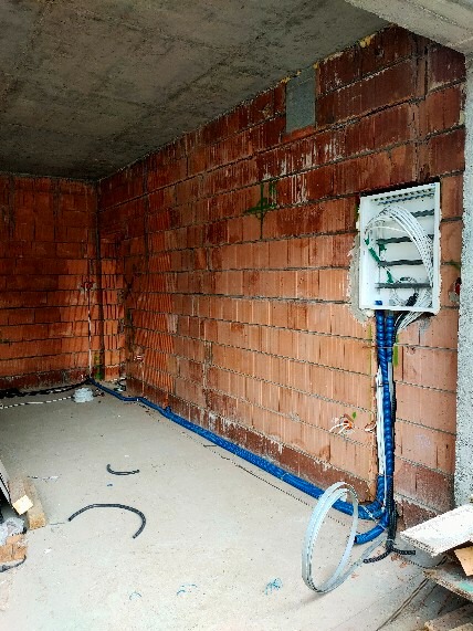 Surowy stan: rozdzielnica elektryczna z niebieskimi peszlami na ceglanej ścianie w trakcie montażu, widoczne przewody i fragmenty podłogi z elementami instalacji.