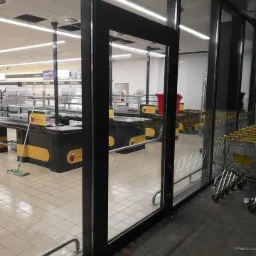 Przez szklane drzwi wejściowe do sklepu widać puste kasy i mop stojący obok jednej z nich. Na zewnątrz, po prawej stronie, rząd wózków sklepowych.